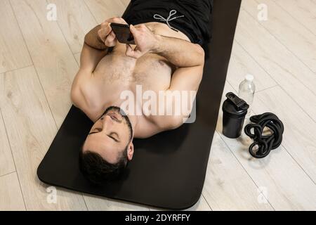 FIT MAN se repose après une forme physique dure et envoie des SMS avec des amis au téléphone. Équipements sportifs. Temps de repos. Paresseux allongé sur un tapis de yoga noir et Banque D'Images