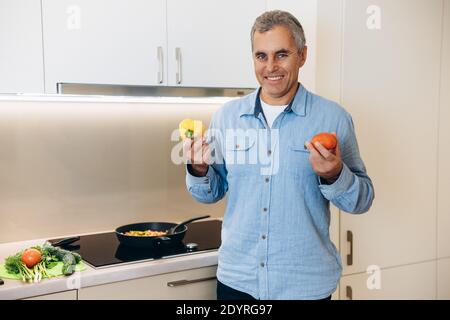 Un homme mûr heureux choisit un légume à ajouter à son plat. Un homme âgé souriant tient du poivre jaune dans une main et de la tomate dans une autre. Cuisine moderne et lumineuse Banque D'Images