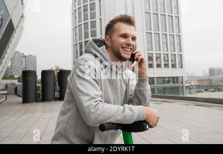 Un homme attrayant avec une barbe raconte à son ami au téléphone la commodité de louer un véhicule électrique. Concept de transport écologique. Décontracté Banque D'Images