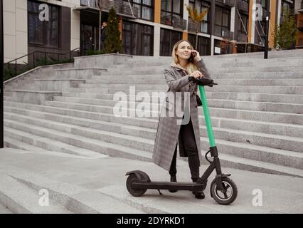 Bonne femme debout près d'un scooter électrique, regardant le ciel et parlant au téléphone avec un ami. Jeune femme en manteau confortable heureux qu'il ya un Banque D'Images