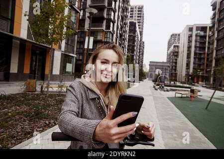 J'aime vraiment cette application de location de scooters femme attrayante en manteau se penche sur le volant d'un scooter, tient un smartphone dans sa main et Banque D'Images