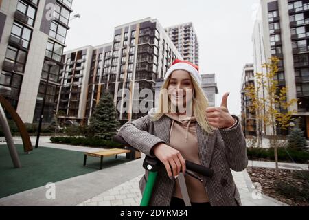 J'aime vraiment ce scooter jeune femme dans le chapeau de Père Noël rouge heureux d'utiliser la nouvelle application de location de scooter de smartphone. Vacances de Noël. Bonne année Banque D'Images