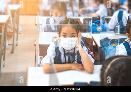 École primaire asiatique en uniforme assise en classe, portant un masque médical pendant l'étude, un écran en plastique sur le bureau de l'étudiant, un étudiant en classe de protection Banque D'Images