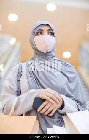 Portrait vertical à la taille de la femme moderne du Moyen-Orient portant un masque et un foulard tout en posant dans un centre commercial Banque D'Images