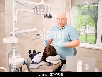 Stomatologue en gants stériles tenant des instruments dentaires et regardant la caméra tout en se tenant à côté du patient. Femme allongé sur une chaise dentaire tout en recevant un traitement stomatologique dans un centre médical Banque D'Images