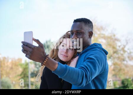 Homme noir africain et femme caucasienne souriant et prenant un selfie avec leur smartphone dans un parc. Portrait de jeune couple multiracial. Banque D'Images