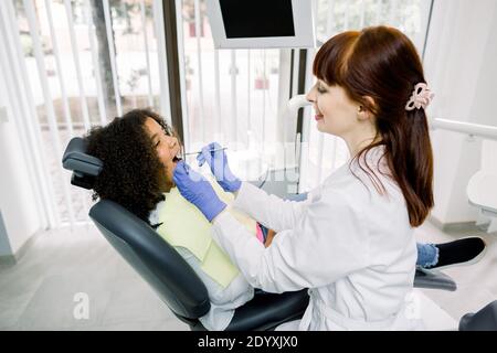 Dentiste et enfant à la clinique dentaire moderne. Jeune dentiste caucasien souriant en uniforme blanc et gants, faisant l'examen de sa petite patiente, peu Banque D'Images