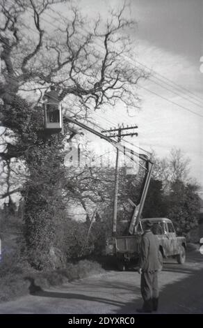 Années 1960, historique, un homme sur un ascenseur aérien à l'arrière d'un Land rover stationné sur une voie de campagne, vérifiant un câble téléphonique aérien coincé dans les branches d'un vieil arbre, Angleterre, Royaume-Uni. Un collègue de travail regardant de la route. Banque D'Images