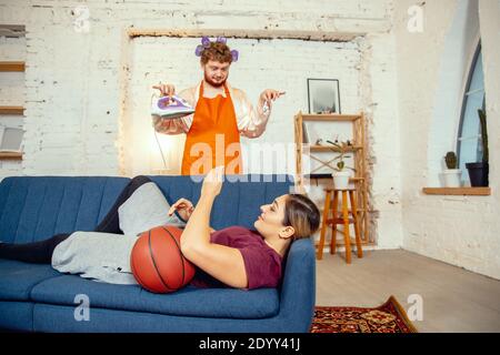 Stéréotypes de genre. Femme et mari faisant des choses inhabituelles pour leurs sexes dans les significations sociales, sens. Homme repassant des vêtements tandis qu'une femme est couché sur le canapé avec ballon de basket dans la salle de séjour. Banque D'Images