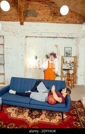 Stéréotypes de genre. Femme et mari faisant des choses inhabituelles pour leurs sexes dans les significations sociales, sens. Homme repassant des vêtements tandis qu'une femme est couché sur le canapé avec ballon de basket dans la salle de séjour. Banque D'Images