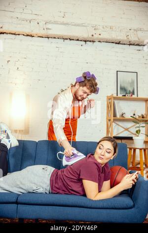 Stéréotypes de genre. Femme et mari faisant des choses inhabituelles pour leurs sexes dans les significations sociales, sens. Homme repassant des vêtements tandis qu'une femme est couché sur le canapé avec ballon de basket dans la salle de séjour. Banque D'Images