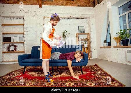 Stéréotypes de genre. Femme et mari faisant des choses inhabituelles pour leurs sexes dans les significations sociales, sens. Homme repassant des vêtements tandis qu'une femme est couché sur le canapé avec ballon de basket dans la salle de séjour. Banque D'Images