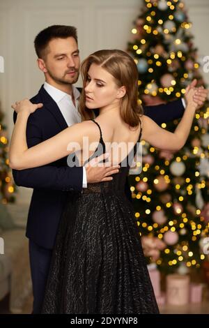 Beau couple dansant dans la salle de séjour. Femme en robe noire et homme en costume se hembrassant en période de Noël. Banque D'Images