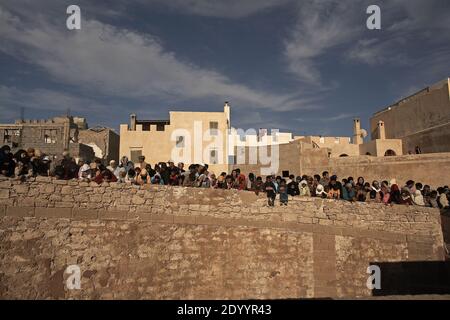 Groupe de personnes assis sur un mur de pierre regardant le Festival mondial de musique de Gnaoua se présentant avec la vieille ville d'Essaouira en arrière-plan. Banque D'Images