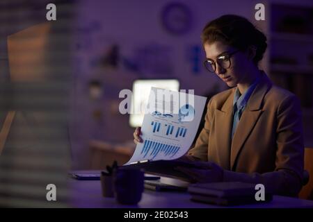 Jeune femme responsable des finances assise à la réception et regardant les journaux tard dans la nuit dans la salle de bureau, espace de photocopie Banque D'Images