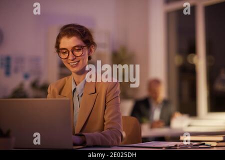 Portrait d'une jeune femme joyeuse portant des lunettes assise au bureau au bureau avec un ordinateur portable tard dans la soirée Banque D'Images