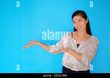 Une prise de vue à la taille d'une femme asiatique attirante dans ses années 30 présentant un produit, pointant avec des mains ouvertes sur un fond bleu clair Uni. Banque D'Images