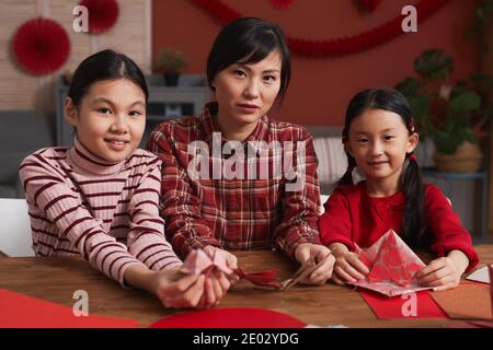 Portrait moyen d'une femme chinoise attrayante avec deux filles assis à une table dans la salle de séjour en train de fabriquer des grues à papier Banque D'Images
