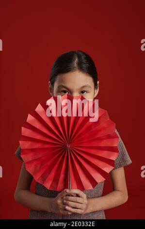 Une fille asiatique méconnaissable se tenant sur un fond de mur rouge tenant traditionnel Ventilateur de papier chinois regardant l'appareil photo Banque D'Images