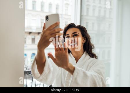 Bonne femme qui prend un selfie sur un balcon dans sa chambre d'hôtel. Femme souriante dans un peignoir tenant un smartphone et faisant des photos sur le devant Banque D'Images
