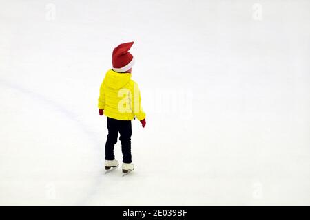 Petit enfant en vêtements d'hiver et casquette rouge du nouvel an se trouve sur la patinoire, vue arrière Banque D'Images