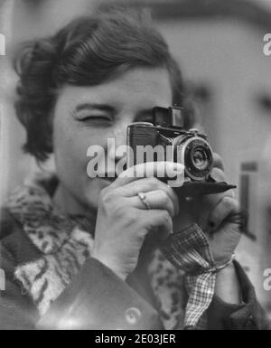 1930s, historique, une jeune femme qui ferme les yeux pour se concentrer alors qu'elle prend une photo avec un appareil-photo compact pliable 'Nagel' Vollenda. Créée par le Dr. August Nagel, co-fondateur de Zeiss Ikon, à Stuggart, en Allemagne, en 1928, la société est devenue connue pour son appareil photo petit format Nagel-Pupille. L'appareil photo utilisé ici est un Vollenda 48, un appareil photo compact pliable qui produit 16 expositions sur film 127. Il avait un viseur optique pliable. Banque D'Images