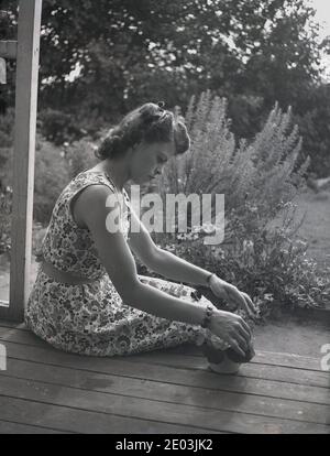 années 1950, historique, une jeune femme portant une robe sans manches à motifs floraux, un article populaire de la mode féminine de cette époque, assise sur une terrasse en bois à l'arrière d'une maison près du jardin, arrangeant des fleurs dans un petit pot. Banque D'Images