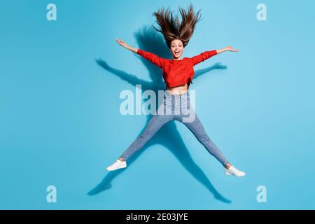 Photo pleine longueur de la folle dame sauter les bras écartés jambes cheveux volant porter rouge chandail baskets jeans isolé bleu couleur de fond Banque D'Images