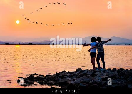 La mère et la fille étaient contents de l'eau qui s'est répandue ses bras pour voler un troupeau d'oiseaux volant une rangée sur le soleil du ciel au coucher du soleil belle nature terre Banque D'Images