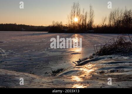 Coucher de soleil orange sur lac glacé glacé réfléchissant avec silhouettes d'arbres sombres, longues ombres et ciel clair Banque D'Images