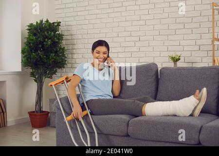 Jeune femme souriante avec jambe cassée assise sur le canapé à à la maison et au téléphone Banque D'Images