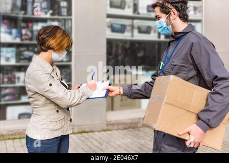 L'homme d'accouchement portant un masque facial et un paquet livré à la femme portant un masque, en gardant la distance sociale pendant que la femme signe l'accouchement Banque D'Images