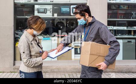 L'homme d'accouchement portant un masque facial et un paquet livré à la femme portant un masque, en gardant la distance sociale pendant que la femme signe l'accouchement Banque D'Images