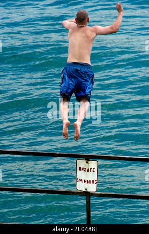 Un homme sautant d'une rampe dans l'eau avec un panneau Dire dangereux pour la natation Banque D'Images