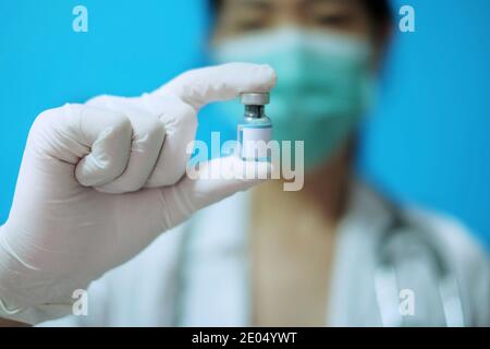 Médecin asiatique féminin avec masque chirurgical et gants en caoutchouc blanc dans une clinique, tenant une bouteille de vaccin en verre avec une étiquette blanche vierge. Banque D'Images