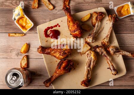 Plat d'une table en bois désordonné avec une assiette de pilons de poulet frits ou grillés avec demi mangé, restes d'os, sauces en immersion i Banque D'Images