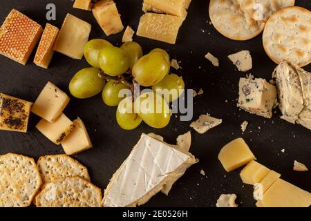 Image plate de l'assortiment de fromages artisanaux français, italiens et suisses vieilli sur plateau de fromages noirs servi avec des craquelins et des raisins. Des crumbles sont visibles ac Banque D'Images