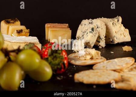 Une sélection de fromages artisanaux français, italiens et suisses sur plateau de fromages noirs servis avec des craquelins et des fruits (raisins et paille) Banque D'Images
