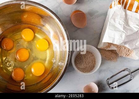 Pâtisserie en cours de préparation sur un comptoir de cuisine en marbre avec un sac en papier de farine de blé entier, des coquilles d'œufs cassées, une tasse à mesurer et un accessoire de mixeur vus. R Banque D'Images