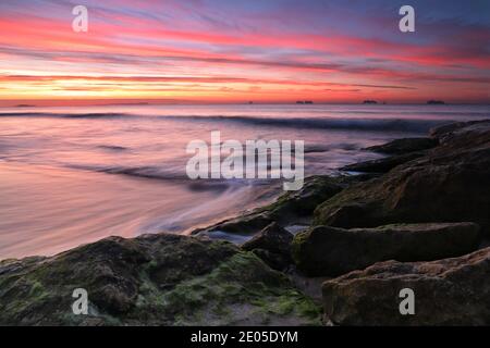 Le soleil se lève dans un ciel parsemé de couleurs chaudes jaune et orange tandis que la mer bat sur les rochers d'une géne de défense de mer en dessous. Banque D'Images