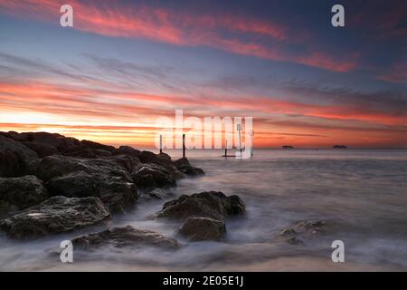 Le soleil se lève dans un ciel parsemé de couleurs chaudes jaune et orange tandis que la mer bat sur les rochers d'une géne de défense de mer en dessous. Banque D'Images