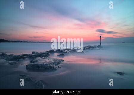 Une longue exposition réduit les vagues de la mer à une texture laiteuse et paisible, tandis que le sable humide reflète les couleurs du soleil d'hiver qui monte. Banque D'Images
