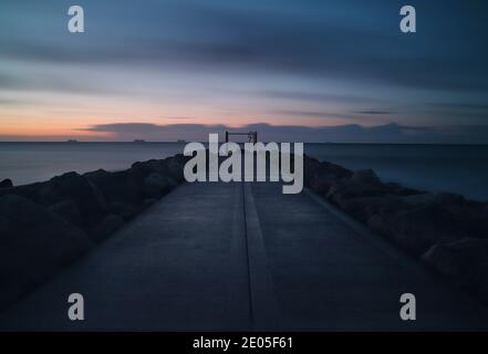 Une longue exposition capture les raindouds gris foncé qui traversent un ciel d'hiver sur un sentier illuminé de gloomies flanqué de rochers qui mènent vers l'horizon. Banque D'Images