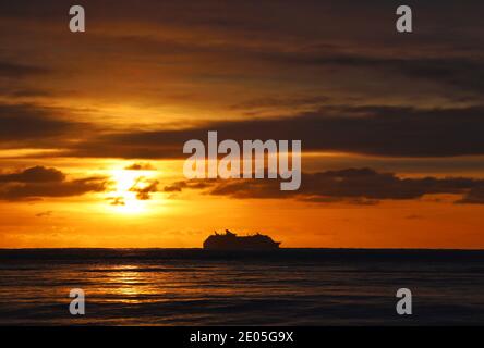 Un bateau de croisière est silhoueté à l'horizon contre un ciel matinal d'hiver rempli de la couleur dorée chaude du soleil levant qui éclate à travers les nuages. Banque D'Images