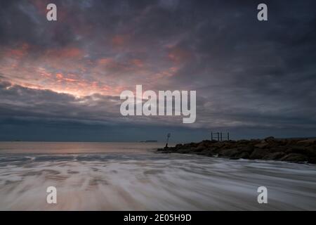 Les vagues se précipitent sur la rive tandis qu'une pointe de couleur rose force son chemin dans un ciel subdué rempli de nuages gris lors d'un matin d'hiver froid. Banque D'Images