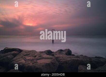 Le soleil commence à se lever dans un ciel rempli de nuages brumeux. Un seul poteau de groyne se lève au centre de l'image hors d'une mer calmé par une longue exposition. Banque D'Images