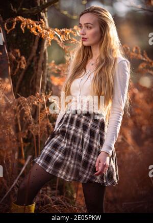 Une jeune belle femme ( 20) dans un cadre de forêt est baignée dans le soleil d'hiver automnal dans une image éditoriale de mode. Porte une jupe tartan et un haut blanc Banque D'Images