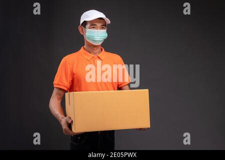 Livraison un homme asiatique ou un coursier portant un uniforme orange masqué tient une boîte en carton dans le studio de concept de coronavirus. Banque D'Images
