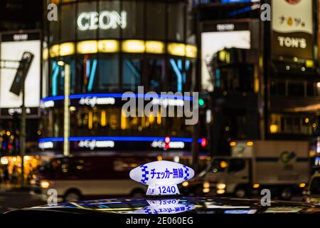 Un taxi portant l'enseigne „Checker Musen“ sur le toit attend au premier plan devant la façade vitrée lumineuse bleue du San-ai Dream Center sur Suzuran-dori à Ginza, Tokyo. Construit en 1963, le bâtiment rond est un repère du quartier. Banque D'Images