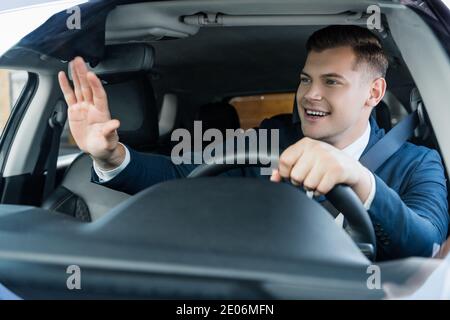 Homme d'affaires gai en agitant la main tout en conduisant la voiture sur un premier plan flou Banque D'Images